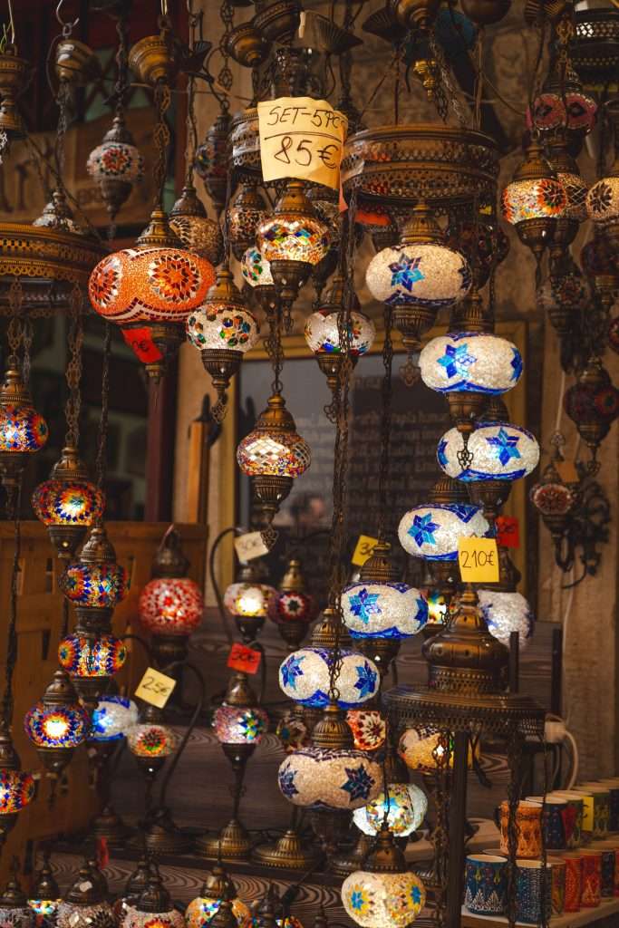 Hanging mosaic lamps with vibrant colors and intricate designs are on display, reminiscent of a magical One Day In Mostar. Paper tags with prices dangle from some lamps, including one marked "SET 5PCS 85€". The lamps boast blue star patterns and textured surfaces.