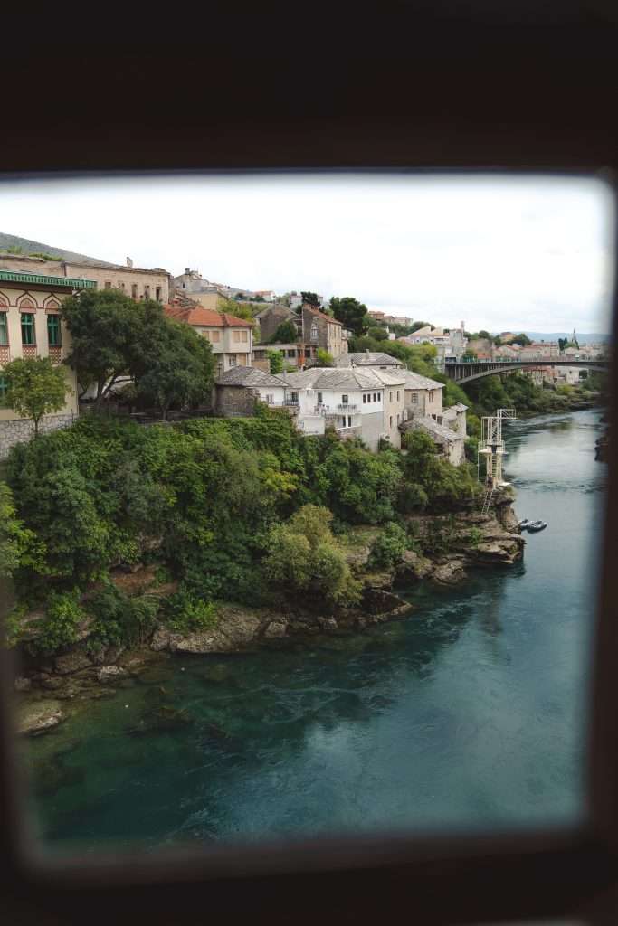 View of a picturesque riverside town with lush greenery and historic buildings atop cliffs, overlooking a calm, flowing river. It's as if you're spending One Day In Mostar. A distant arched bridge spans the water under a cloudy sky, with the image framed by a dark border.