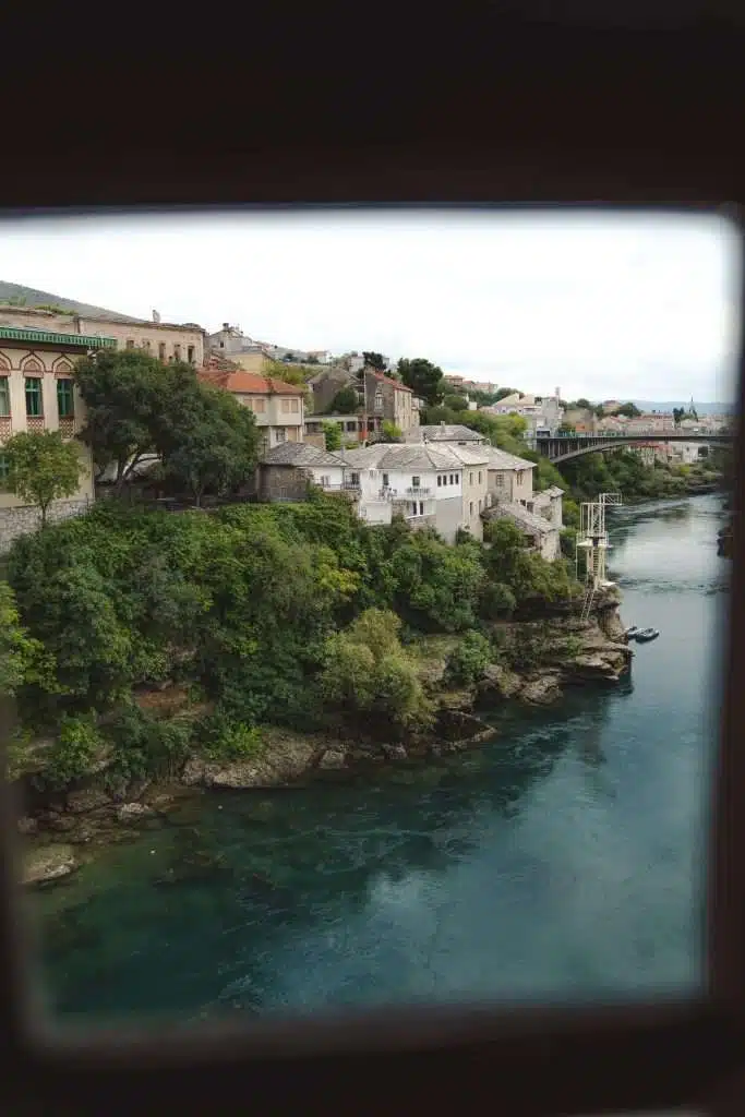 View of a picturesque riverside town with lush greenery and historic buildings atop cliffs, overlooking a calm, flowing river. It's as if you're spending One Day In Mostar. A distant arched bridge spans the water under a cloudy sky, with the image framed by a dark border.