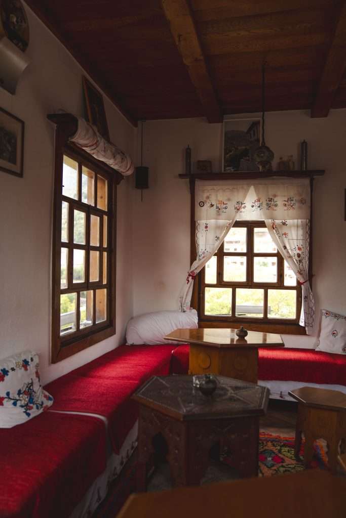 Cozy room with a wooden ceiling, windows allowing natural light to illuminate red cushioned benches and a small wooden table. Embroidered curtains and a patterned carpet add a traditional touch, reminiscent of spending One Day In Mostar.