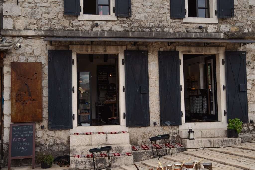 A rustic stone building in Mostar features two open doors and black shutters. A wooden sign on the left displays wine options, reminiscent of a one-day visit. A paved stone path leads to the entrance, with green potted plants adorning the facade.