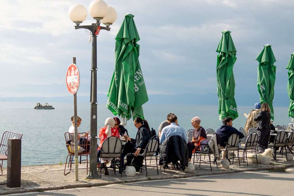 People sit at outdoor café tables by a waterfront, under green umbrellas. A lamppost and a sign are nearby. In the background, a body of water and a boat are visible under a cloudy sky—a scene straight out of your One-Day in Lake Ohrid itinerary.