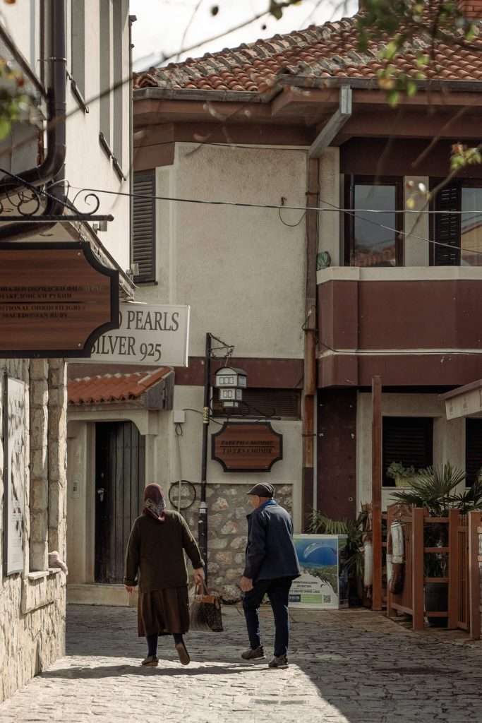 A couple strolls down a narrow cobblestone street, enveloped by charming buildings with signs like "Silver 925." Trees with sparse leaves and wooden-accented, tile-roofed architecture set a picturesque scene reminiscent of a one-day in Lake Ohrid itinerary.