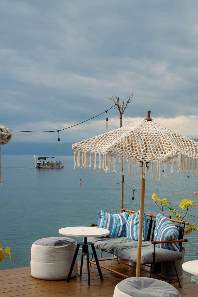 Cozy outdoor seating area by a tranquil lake, perfect for a One-Day In Lake Ohrid Itinerary. A white fringed umbrella shades a cushioned sofa with striped pillows. Under the cloudy sky, a boat drifts on the water while string lights add charm to the serene scene.