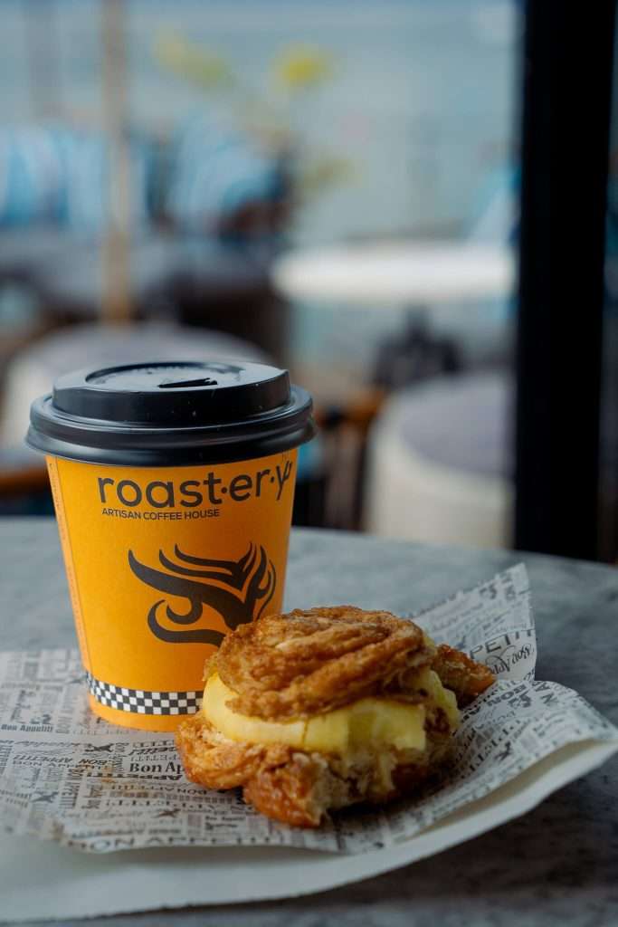 A yellow coffee cup with a black lid labeled "roastery" sits on a table, evoking memories of a perfect One-Day In Lake Ohrid Itinerary. Next to it, a pastry filled with cream rests on newspaper-style paper, all set against the softly blurred backdrop of a cozy café.