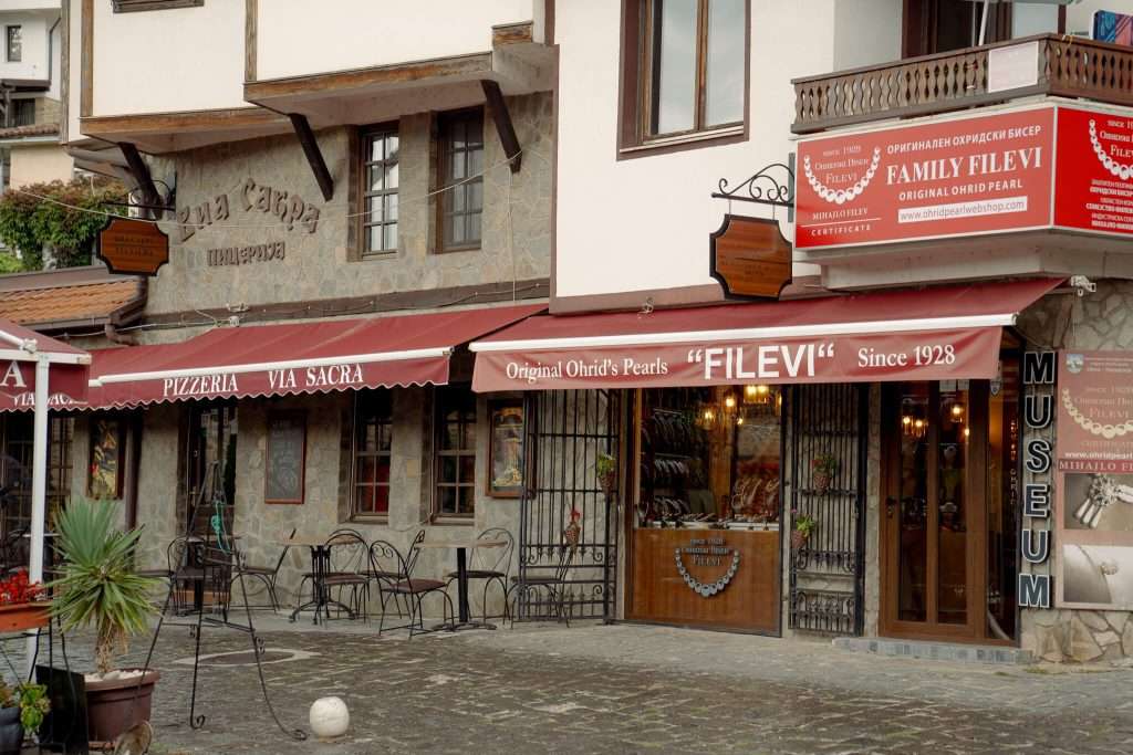 A European-style building offers a charming street view in Lake Ohrid. It houses a pizzeria and a store selling Ohrid's pearls, with stone walls, red canopies, and outdoor seating. Part of your One-Day In Lake Ohrid Itinerary, it sits near a "Museum" sign gracefully displayed on the side.