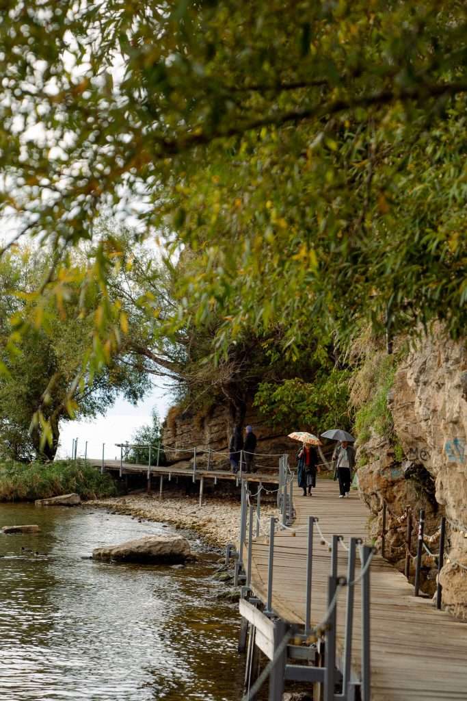 A scenic lakeside path, perfect for a one-day in Lake Ohrid itinerary, where people walk under umbrellas. Flanked by rocky cliffs on the right and overhanging trees on the left, it overlooks a calm lake. The overcast sky hints at a gentle drizzle, adding to the atmosphere.