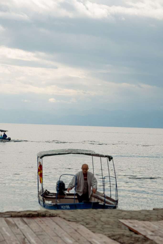 A man stands on a small boat with a canopy on the calm waters of Lake Ohrid. Another boat traces its path in the distance, softly slicing through the serene expanse. The sky is overcast, with clouds gently diffusing light, setting a tranquil stage for your one-day Lake Ohrid itinerary.