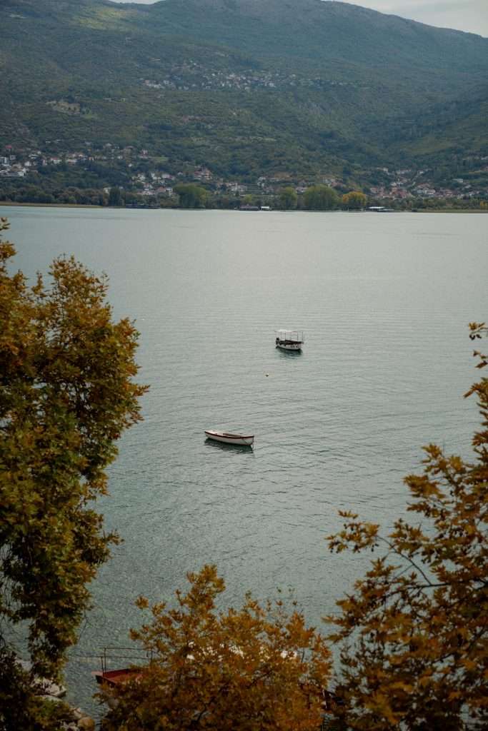 A peaceful lake scene with two small boats floating on calm water, much like a serene moment in a One-Day In Lake Ohrid Itinerary. Surrounding the lake are lush, tree-covered hills under a cloudy sky, while autumn leaves frame the foreground.