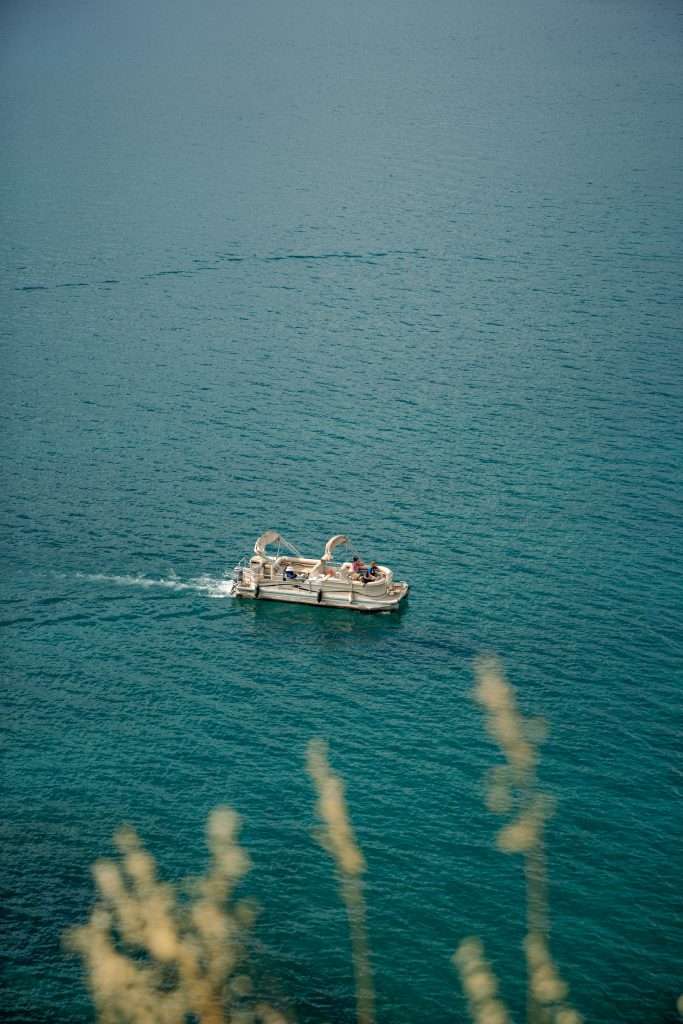 A small boat with passengers sails on a calm, deep blue sea as part of a One-Day In Lake Ohrid Itinerary. The foreground features blurred golden reeds, adding depth to the serene ocean scene.