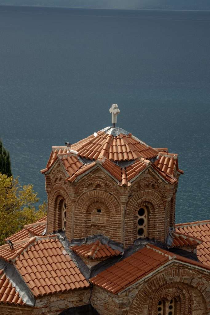 Nestled by the tranquil waters, a historic brick church with a cross-topped red-tiled roof showcases arched windows, offering a serene stop in your One-Day In Lake Ohrid Itinerary.