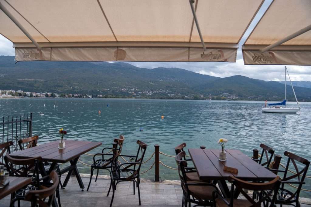 Outdoor patio with wooden tables and chairs overlooking a tranquil lake, perfect for your One-Day In Lake Ohrid Itinerary. A sailboat floats on the water, with mountains and a small town in the background. An awning partially covers the seating area.