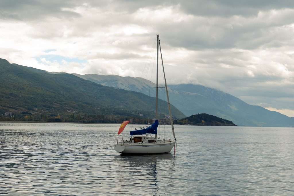 A sailboat with a blue cover and a small flag floats on the serene Lake Ohrid, its calm waters framed by mountains under a cloudy sky. The distant shoreline, part of your one-day itinerary, is adorned with lush greenery.