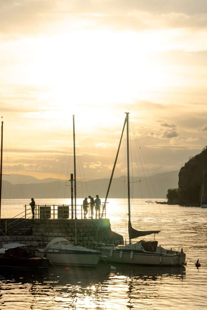 As the sun sets over Lake Ohrid, a group of people stands silhouetted on a stone pier, savoring the end of their one-day itinerary. Sailboats gently bob nearby, their reflections dancing in the golden water. In the distance, hills bask under a partly cloudy sky.