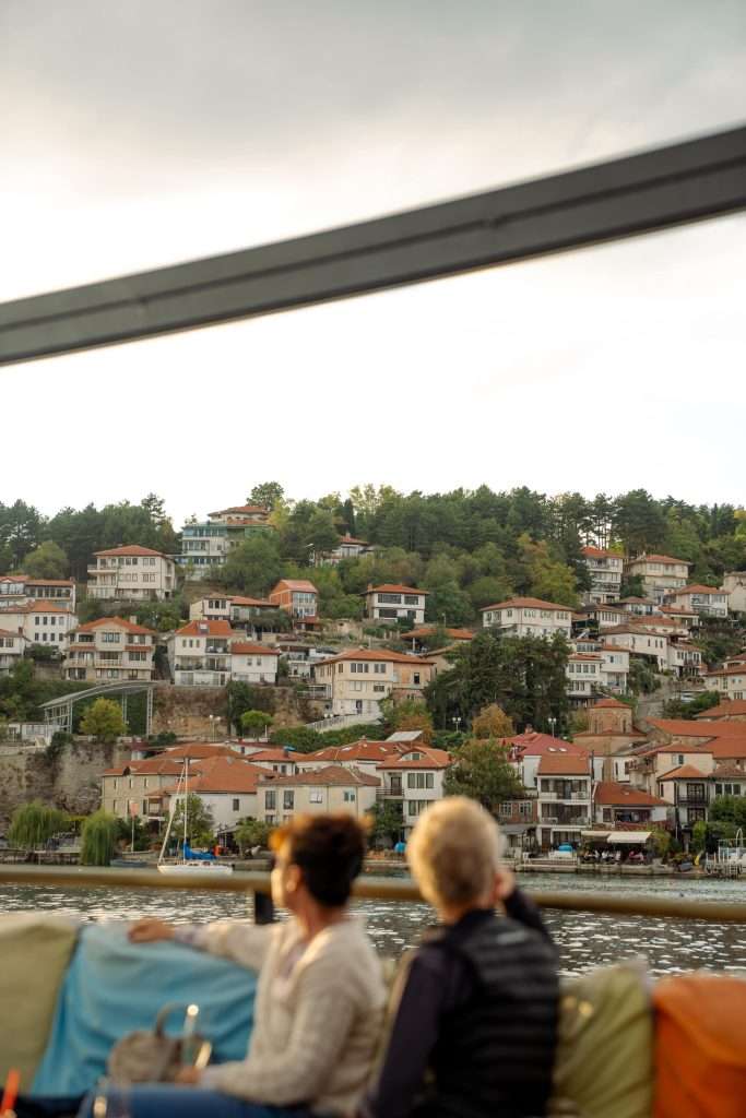 A scenic view of a hillside town with terracotta-roofed houses surrounded by lush greenery offers a perfect backdrop for your One-Day in Lake Ohrid Itinerary. Two people stand in the foreground, gazing across the glistening water toward the charming town as a sailboat drifts near the shore.