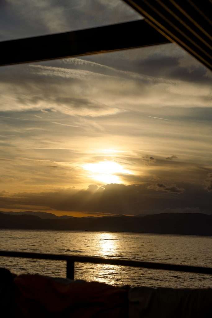 Sunset over Lake Ohrid, viewed through a railing. The sky is painted in hues of orange and gold as the sun sets behind wispy clouds, reflecting off the calm water. A serene and picturesque scene, perfect for ending a One-Day In Lake Ohrid Itinerary amidst nature's splendor.