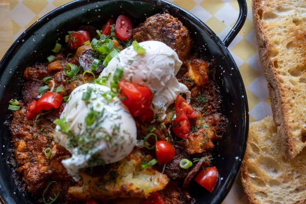 On a patterned tablecloth reminiscent of a quaint café, a skillet brims with poached eggs, diced tomatoes, green onions, and herbs over savory potatoes and sauce. It’s the kind of hearty meal you dream of after just one day in Sofia. Slices of toasted bread eagerly await beside it. Dish is the Potato Pan from Cafe 1920 - Sofia, Bulgaria