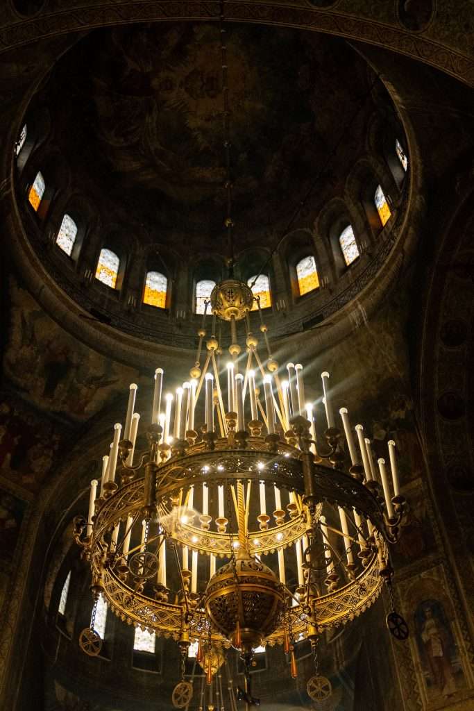 A grand chandelier with numerous candle-like lights hangs from the ornate dome of a church (Cathedral of St. Alexander Nevsky) , casting a soft glow on this serene stop during one day in Sofia. The dimly lit interior highlights intricate designs, stained glass windows, and detailed artwork adorning the walls and ceiling.