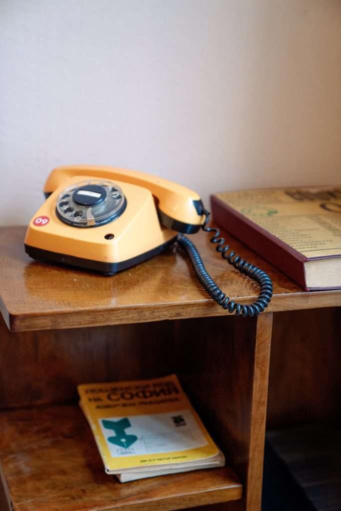 A vintage orange rotary telephone rests on a wooden shelf next to a closed book and a yellow magazine, reminiscent of One Day In Sofia. The phone's cord is coiled and draped casually, evoking a nostalgic atmosphere.