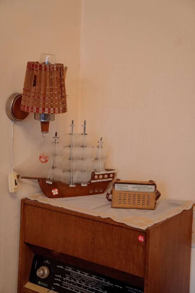 In a vintage wooden cabinet from One Day In Sofia, an old radio sits beside a model ship with white sails and small red emblems. A woven wall lamp with a red shade casts a soft glow, setting the scene in a warmly lit, beige room.