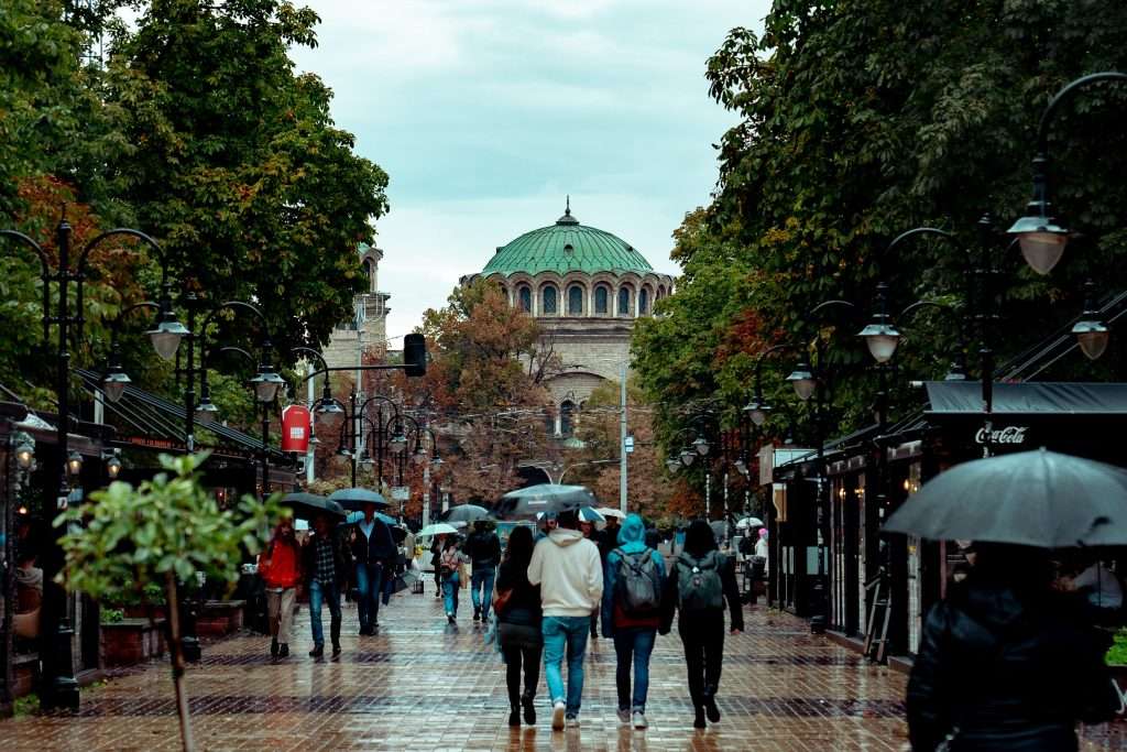 On a rainy day in Sofia on Vitosha Boulevard , people with umbrellas stroll down a wet, tree-lined street. Shops and lampposts line the sides, while an ornate building capped with a green dome stands under the cloudy sky in the background.