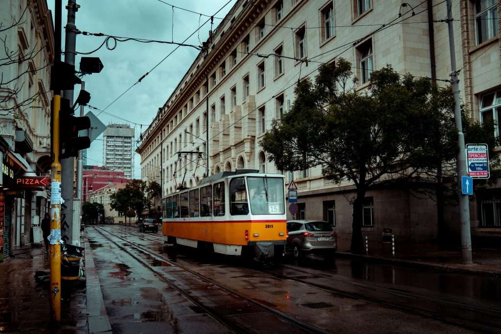 A yellow and white tram travels down a wet street in Sofia, lined with tall, historic buildings on a rainy day. The road glistens from the rain, with a few cars and trees visible along the side. It captures the essence of an urban cityscape under overcast skies duing 2 days in Sofia.