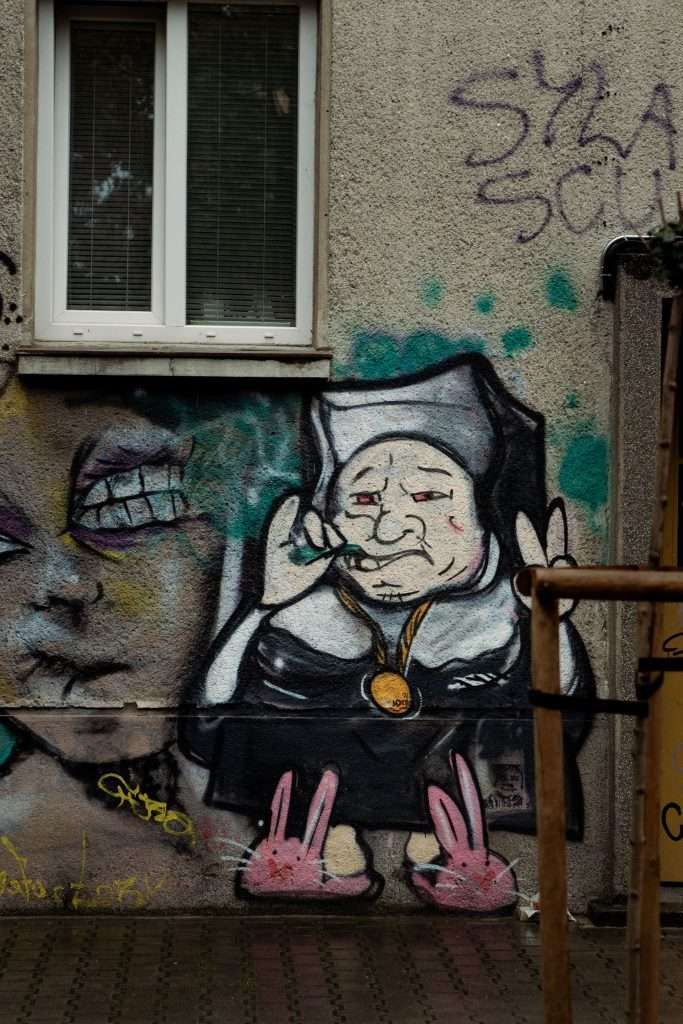 Graffiti on a wall whimsically captures the scene of a nun in pink bunny slippers, glasses perched on her nose, and a playful expression. Beside her, colorful abstract artwork unfolds vibrantly, reminiscent of One Day In Sofia. A window peeks into this lively tableau from above.