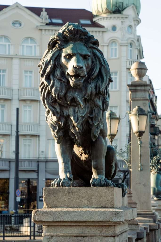 A bronze lion statue sits majestically on a stone pedestal in front of an ornate building in Sofia. This sculpture is part of a bridge adorned with vintage-style streetlamps, creating a charming scene bathed in soft daylight—a perfect snapshot for One Day In Sofia.