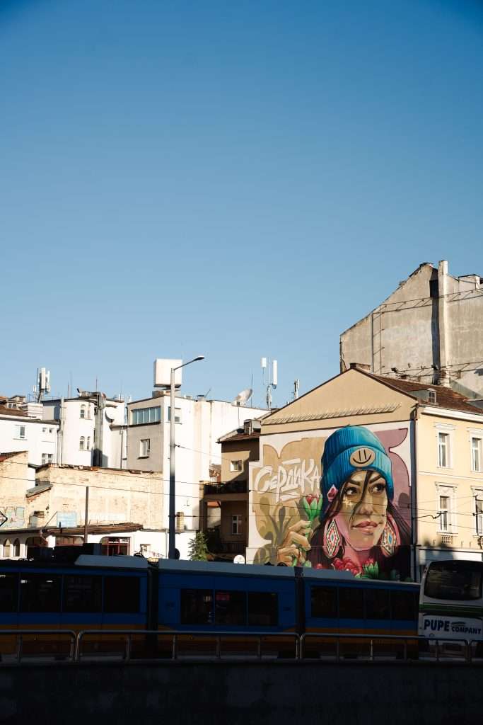 A colorful mural of a woman wearing a blue hat graces a building in an urban setting. It's "One Day In Sofia," with a clear blue sky above. Below, part of a streetcar or tram passes by, capturing the vibrant life of the city.