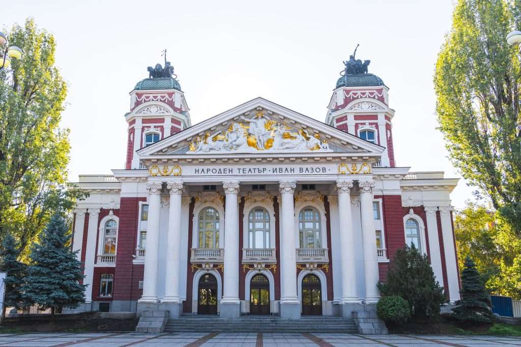 One Day In Sofia reveals a majestic neoclassical building adorned with grand columns and ornate architectural details, gracefully surrounded by trees. Its facade boasts intricate sculptures and Cyrillic text, capturing the essence of Sofia's rich cultural tapestry. City Garden & National Theater "Ivan Vazov"