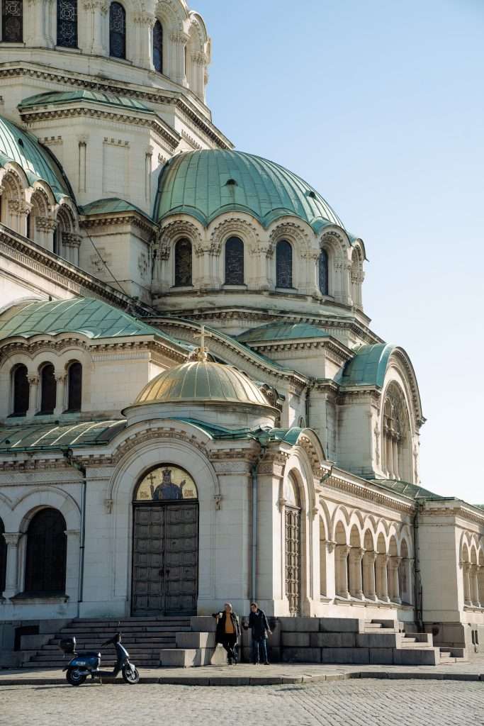 A large, ornate building with multiple domes and arches under a clear blue sky captures the essence of "One Day In Sofia." Two people stand near a door, while a parked motorcycle rests on the cobblestone road in the foreground, offering a glimpse into everyday life.