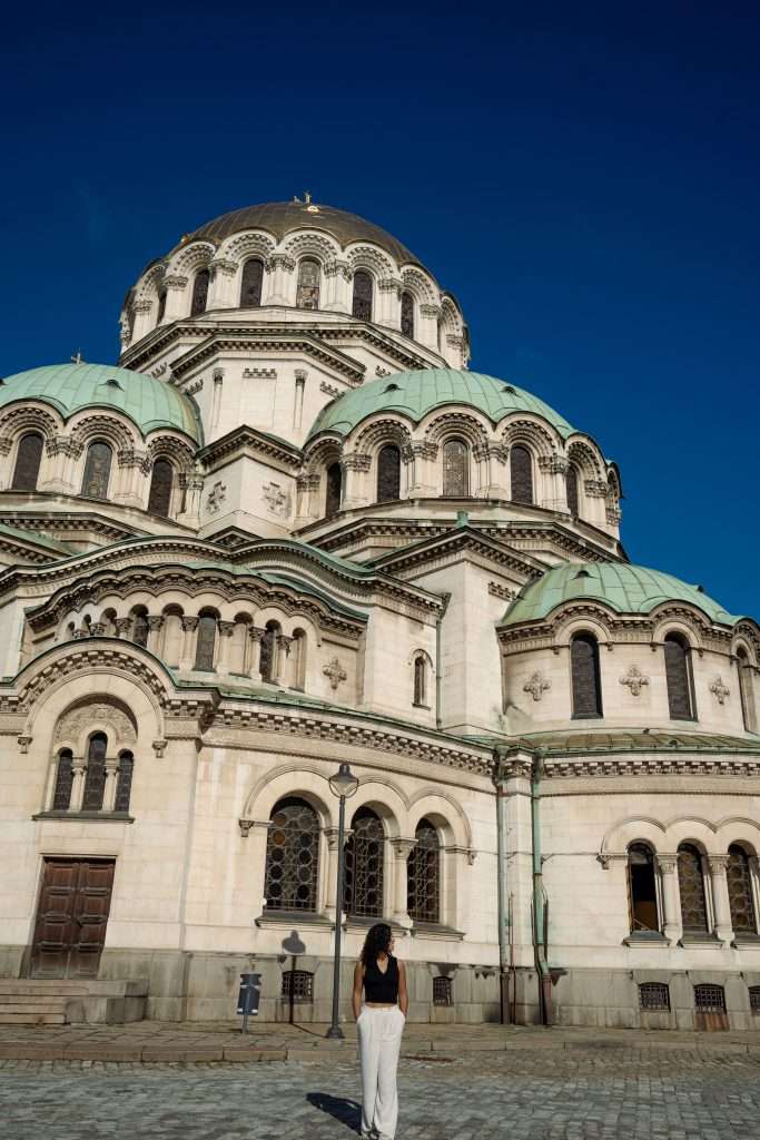 A person stands in front of a large, ornate building (Cathedral of St. Alexander Nevsky) with multiple domes and arched windows against Sofia's clear blue sky. The structure features intricate architectural details, telling tales of One Day In Sofia, with a cobblestone pavement spreading elegantly in the foreground.