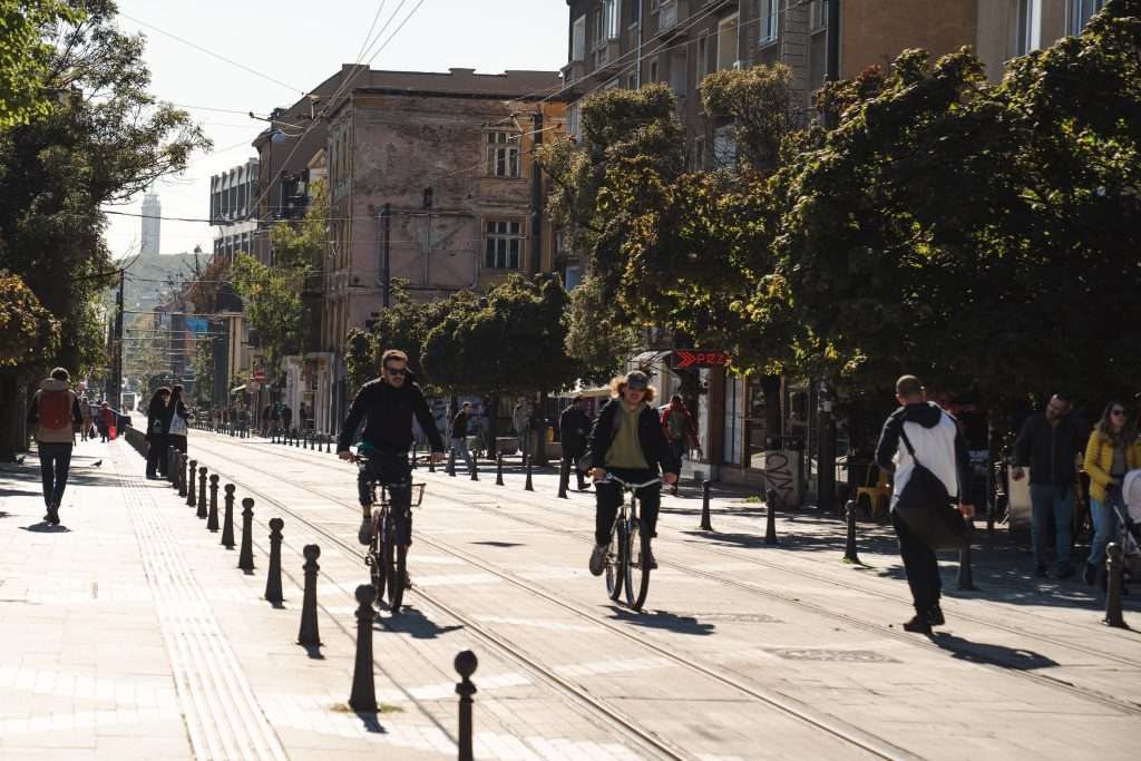 A sunlit urban street in Sofia features tram tracks at its center. People are bicycling and walking along the road. Trees line the sidewalks, with a mix of old and modern buildings flanking the scene. The atmosphere is vibrant and casual, capturing just another day in this charming city.