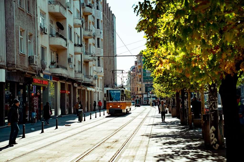 A city street in Sofia bustles with life, as a tram glides along its tracks surrounded by vibrant buildings and trees. Pedestrians stroll the sidewalks, while the sun casts playful shadows. The atmosphere captures the lively yet relaxed essence of spending one day in this charming locale.