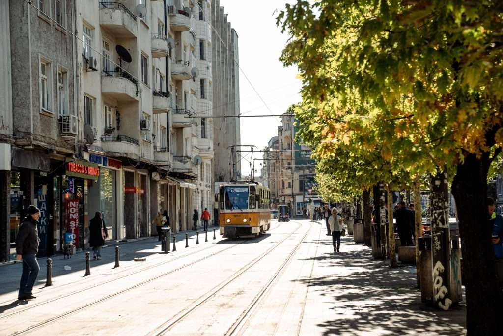 A street scene in Sofia features a yellow tram gliding through the city, flanked by tall buildings on the left. People stroll along sidewalks while leafy trees cast shadows to the right. The sun shines brightly, encapsulating a lively urban vibe as if capturing One Day In Sofia.