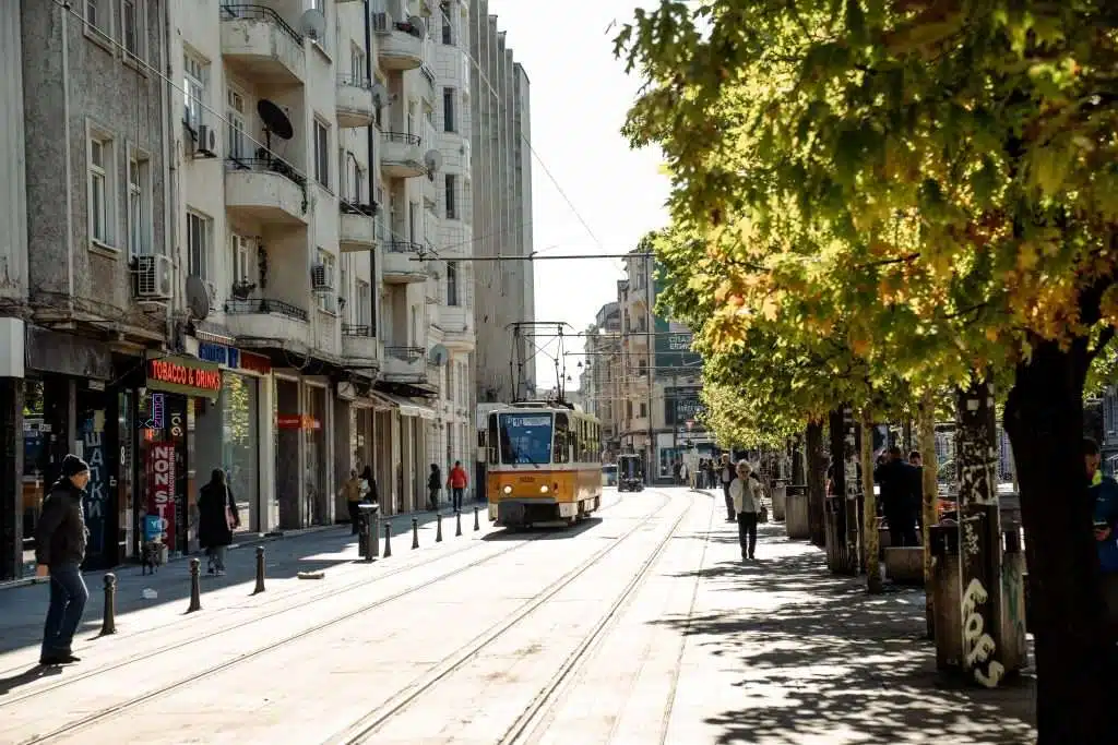 A street scene in Sofia features a yellow tram gliding through the city, flanked by tall buildings on the left. People stroll along sidewalks while leafy trees cast shadows to the right. The sun shines brightly, encapsulating a lively urban vibe as if capturing One Day In Sofia.
