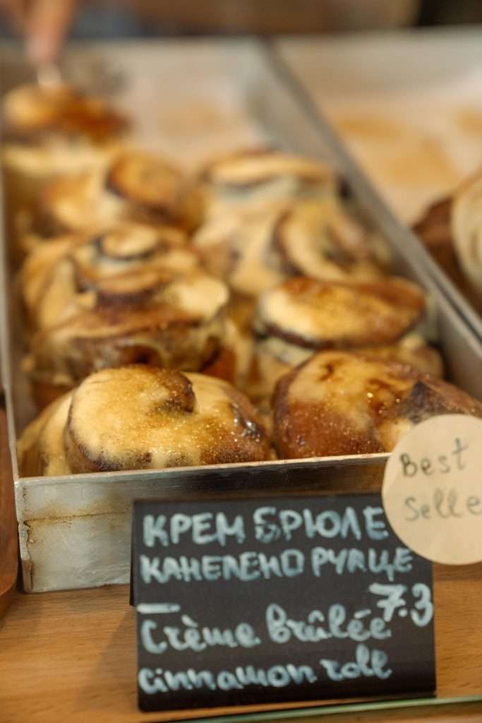 A tray of crème brûlée cinnamon rolls at 369 Bakery is artfully displayed in a bustling bakery, each topped with a caramelized glaze. A sign in front reads, "Crème brûlée cinnamon roll," featuring Russian script and highlighting it as a bestseller—a perfect treat for One Day In Sofia.