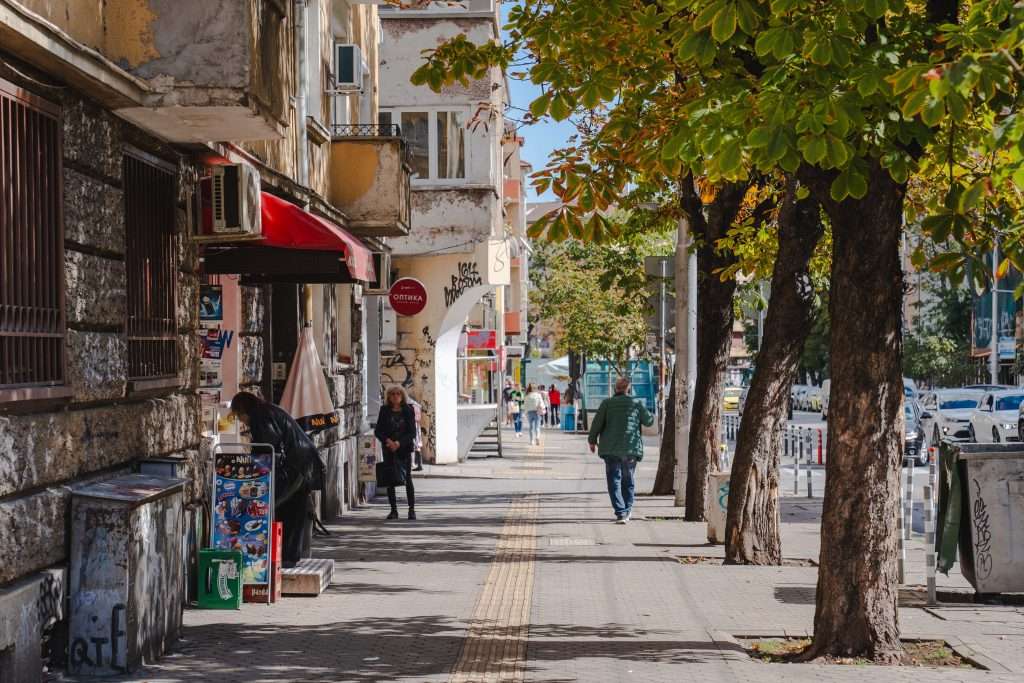 A street scene in Sofia with people walking on a sidewalk lined with trees and buildings. A man in a green jacket walks away, while others stand near the shops. The trees have green leaves, and some walls are covered with graffiti. It's a bright, sunny day, one typical of a vibrant city like Sofia.