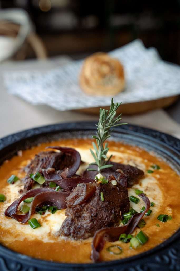 A black bowl brimming with rich, meaty stew featuring tender beef chunks, garnished with green onions and a sprig of rosemary—a taste reminiscent of savoring One Day In Sofia. In the background, a bread roll rests invitingly in a basket.