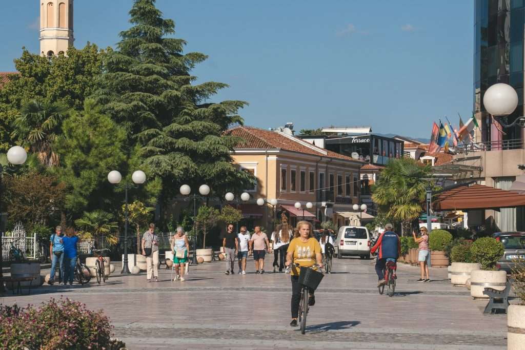 A sunny street scene with people strolling and biking along a wide, paved path. A cyclist wearing a yellow shirt is in the foreground. Under a clear blue sky, trees, flags, and a tall building grace the background.