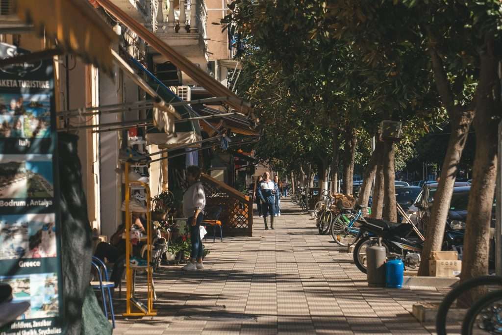 A sunny street scene unfolds as people stroll down a tree-lined sidewalk, reminiscent of a 10-day Albania itinerary. Outdoor cafes with tables and chairs grace the left, bicycles rest along the curb, and parked cars neatly line the street.