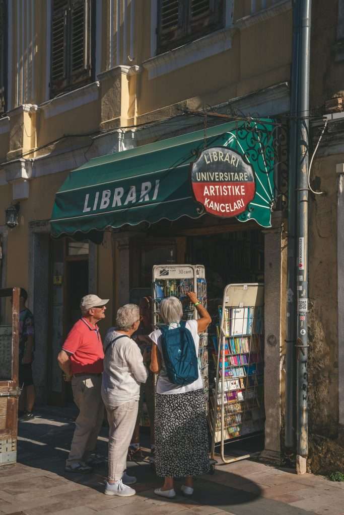 A group of elderly people stands outside a small bookstore with a green awning labeled "Librari," perusing books and postcards  On this sunny day, the building's rustic charm, complete with window shutters, adds to the quaint allure of their journey.