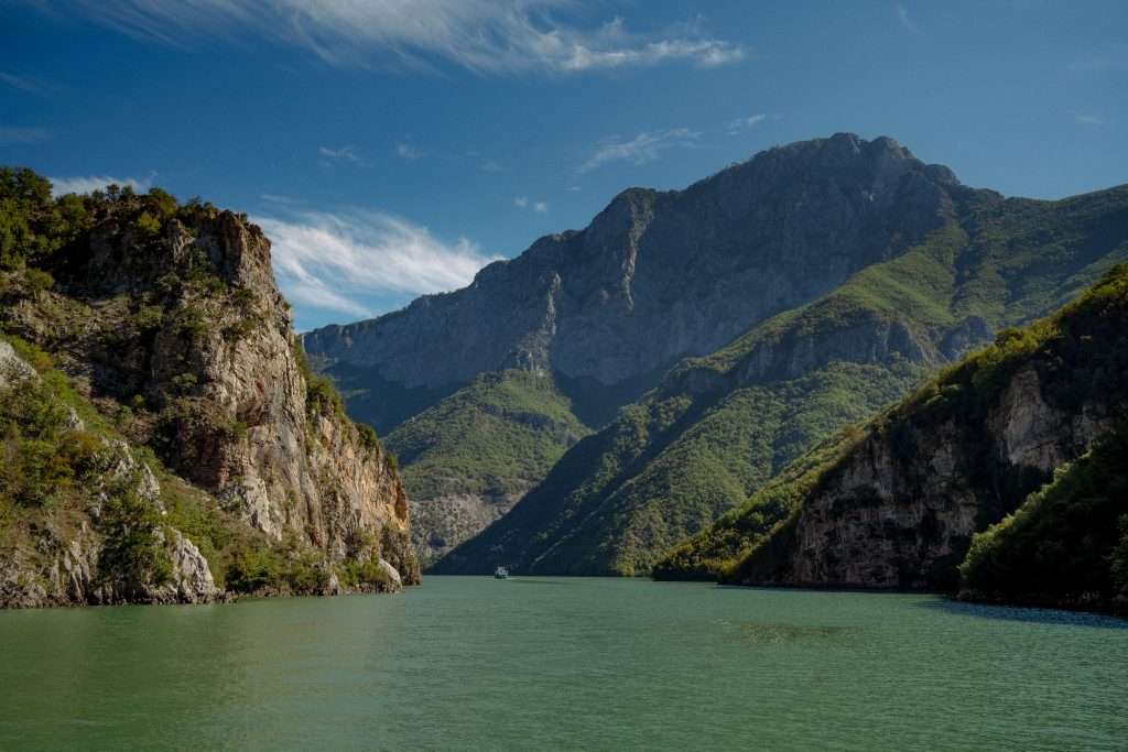 A scenic view of a Koman Lake In Albania flows between two rocky, green hills under a clear blue sky, perfect for your 10-Day Albania itinerary. A small boat is visible in the distance, nestled within the majestic landscape as tall mountains rise in the background.