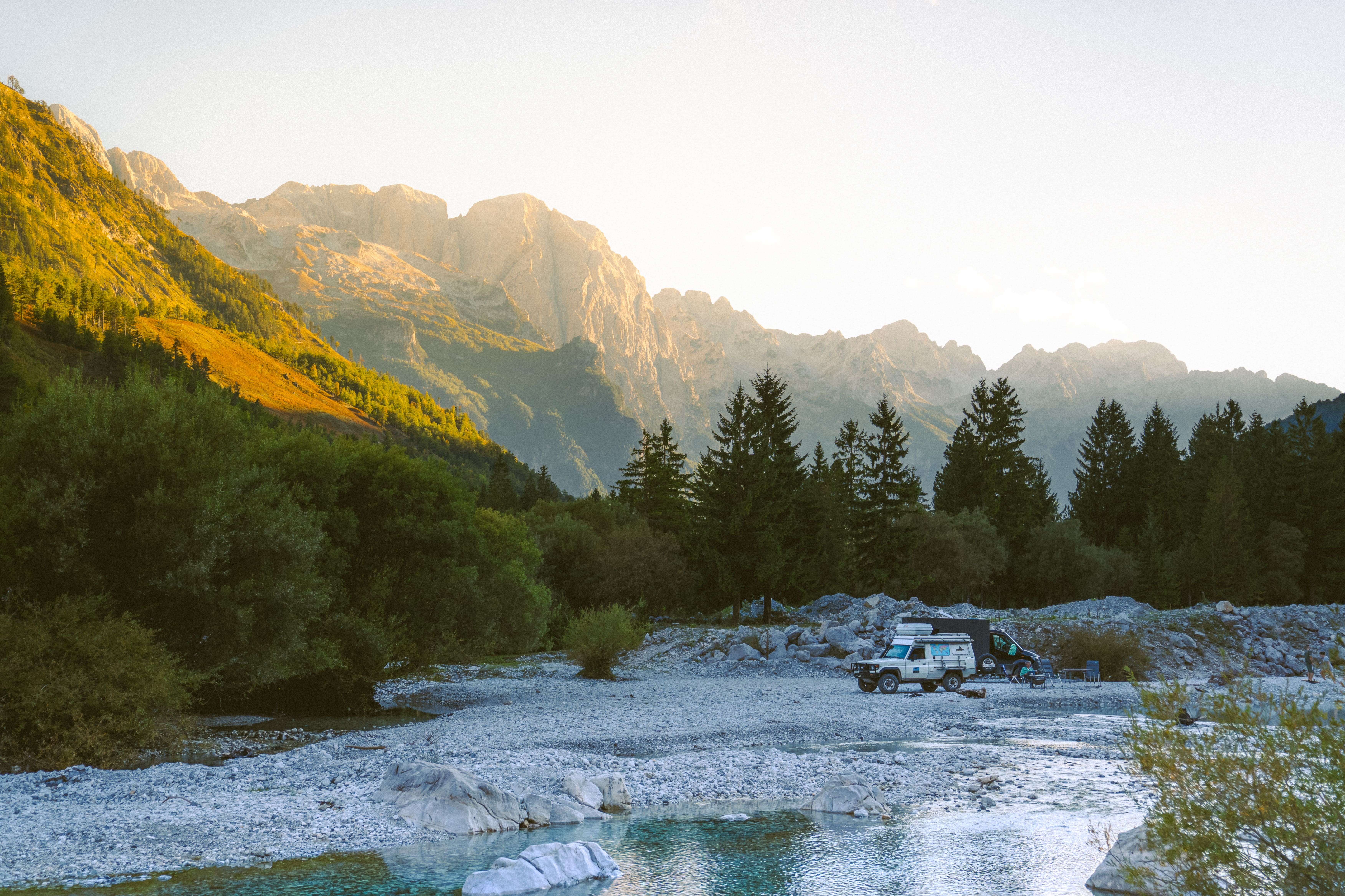 A camper van parked near a river in the picturesque mountains of Valbona in Albania during sunset is perfect for your 10-day itinerary. Tall pine trees and rocky terrain envelop the scene, as sunlight gracefully illuminates the distant peaks, creating an idyllic backdrop for exploration.