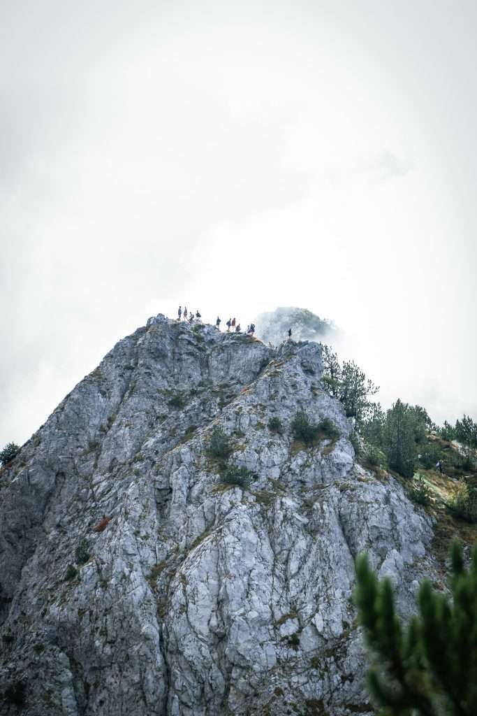 A group of hikers stands atop a rugged, rocky mountain peak, surrounded by mist and scattered greenery. The cloudy sky adds a dramatic atmosphere to the scene, offering a breathtaking highlight to their 10-day Albania itinerary.