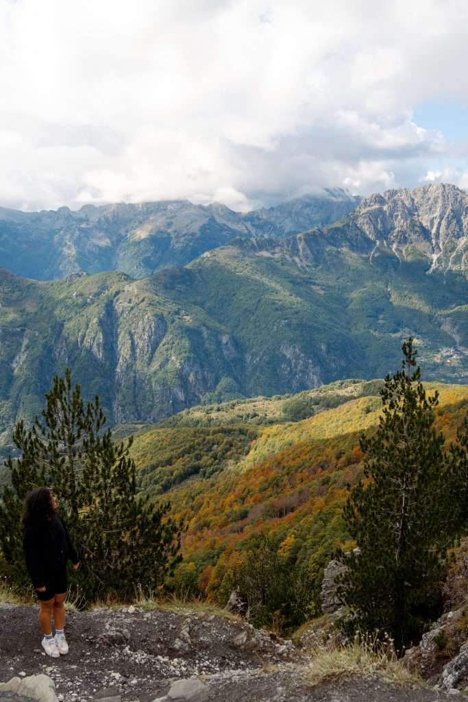 A person stands on a rocky outcrop, gazing at the vast mountainous landscape of their 10-Day Albania Itinerary. The scene features lush green and autumn-colored trees, steep rocky cliffs, and a partly cloudy sky, creating a tranquil and majestic view. Valbona to Theth hike in Albania