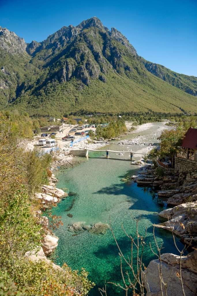 A clear turquoise river winds through a lush Albanian valley with a rocky shore, embraced by green mountains under a crystal blue sky. A quaint footbridge spans the water, while distant buildings hint at charming stops on your 10-Day Albania Itinerary.