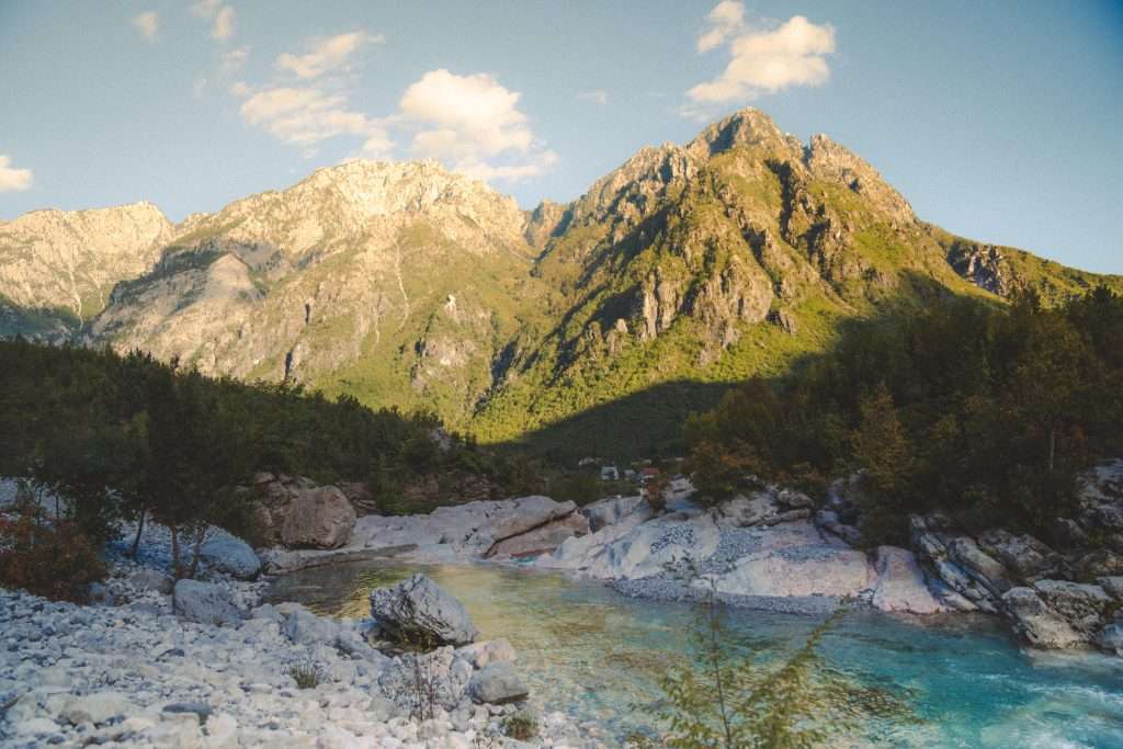 Discover a serene landscape reminiscent of a 10-Day Albania Itinerary, with rugged mountains bathed in warm sunlight, and a clear blue river flowing in the foreground. Lush green trees dot the scene, set against a backdrop of a partly cloudy sky.