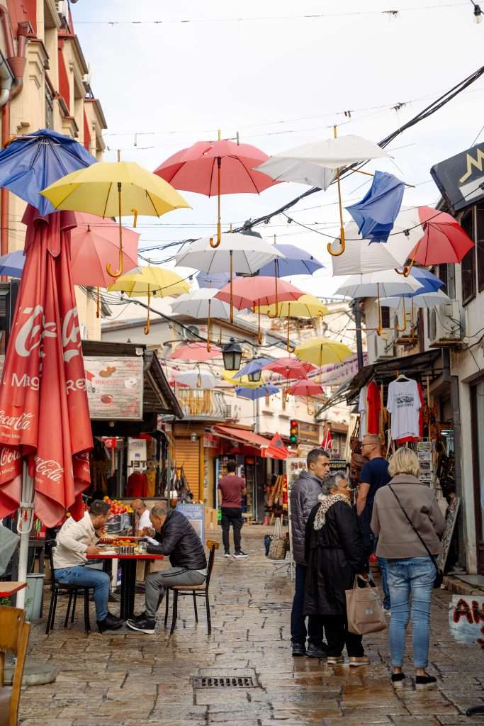 A vibrant street scene in Skopje Old Town Bazaar featuring colourful umbrellas hanging overhead. People walk and sit at cafes, surrounded by shops with various goods on display. The cobblestone pathway adds charm to the lively atmosphere.