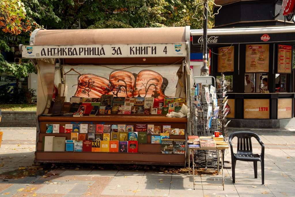 A small outdoor bookstall in Skopje, North Macedonia with a variety of books displayed on shelves and a wire rack sits invitingly. The sign above, in Cyrillic script, with trees and a café in the background offering charm.